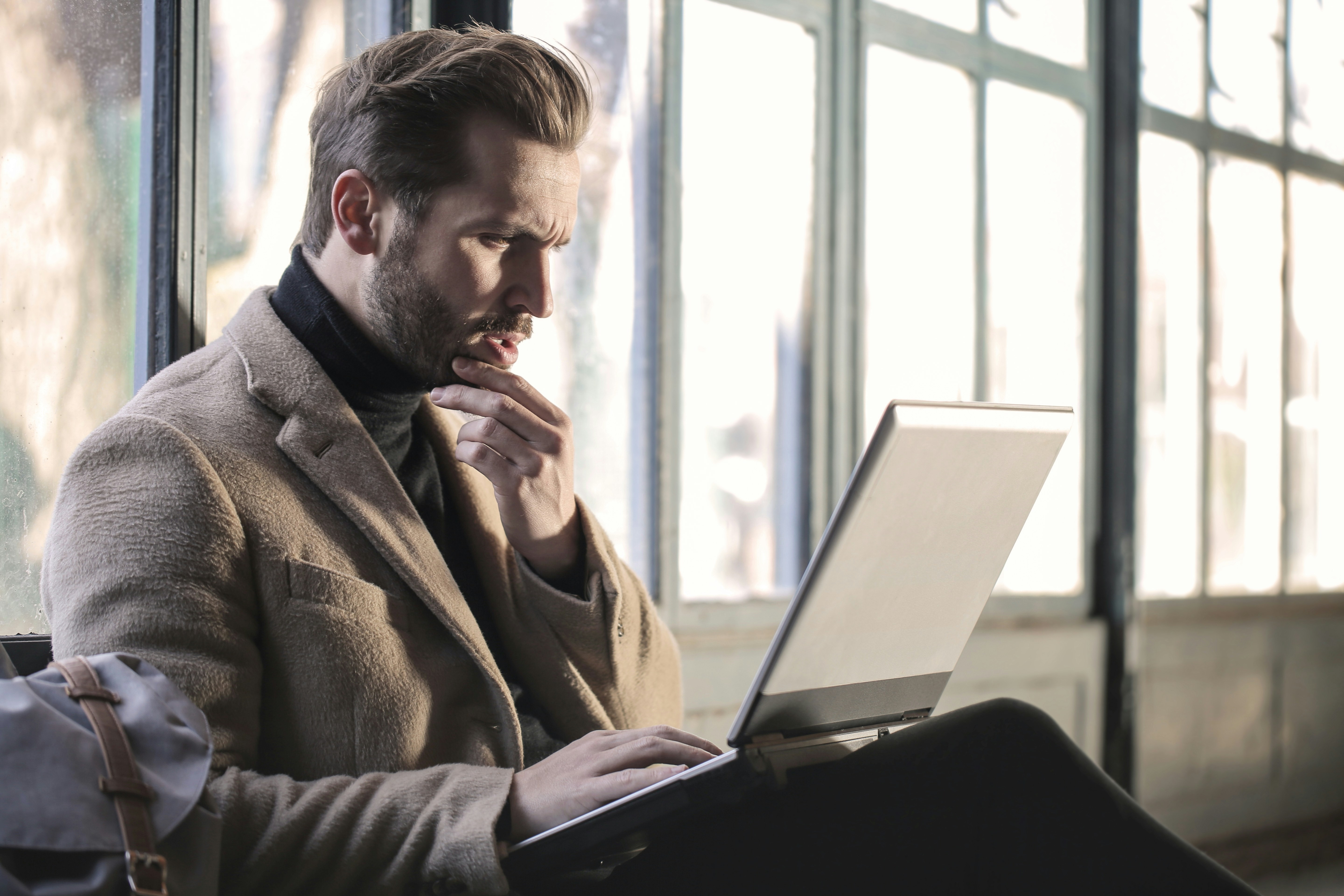 Überraschter Mann in einem Mantel sitzt auf einer Bank und arbeitet an seinem Laptop.